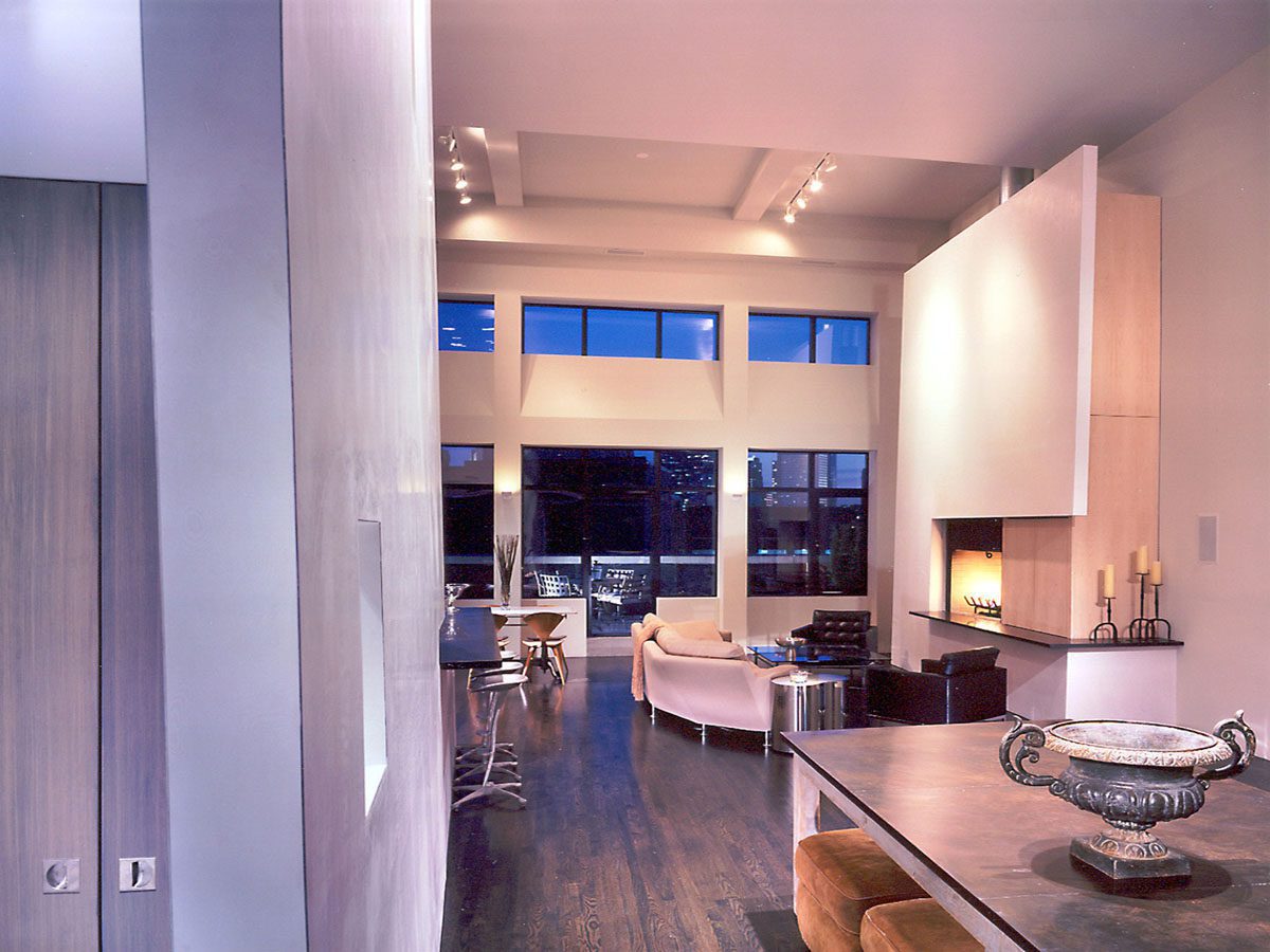 Interior of a loft in Boston, MA, designed by Ruhl Jahnes, showing the open living area with a double-height wall, dark wood floors, a fireplace, low seating, floor-to-ceiling terrace windows, and track lighting above.