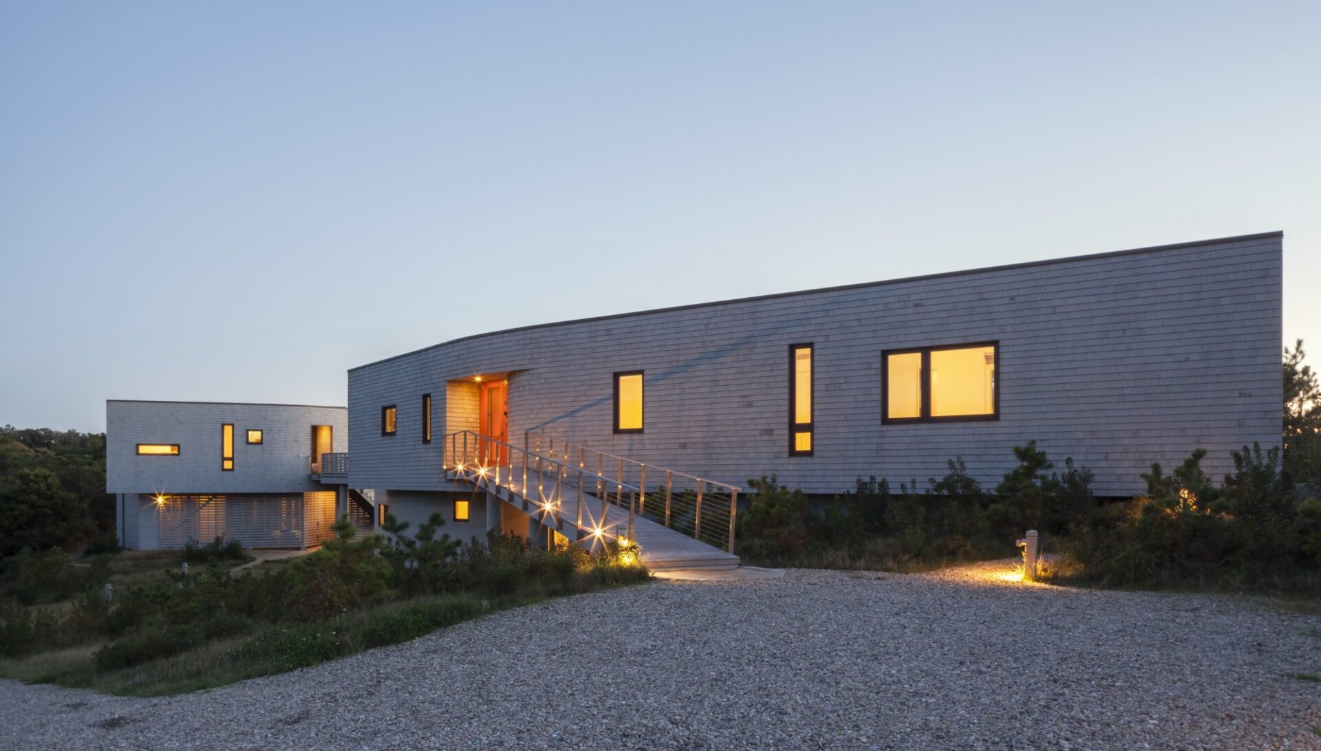 Exterior of a net-zero beach house on Cape Cod, MA, designed by Ruhl Jahnes, showing the curving cedar shingle facade at dusk with glowing windows, a lit accessible entry ramp, and native scrub pine landscaping.