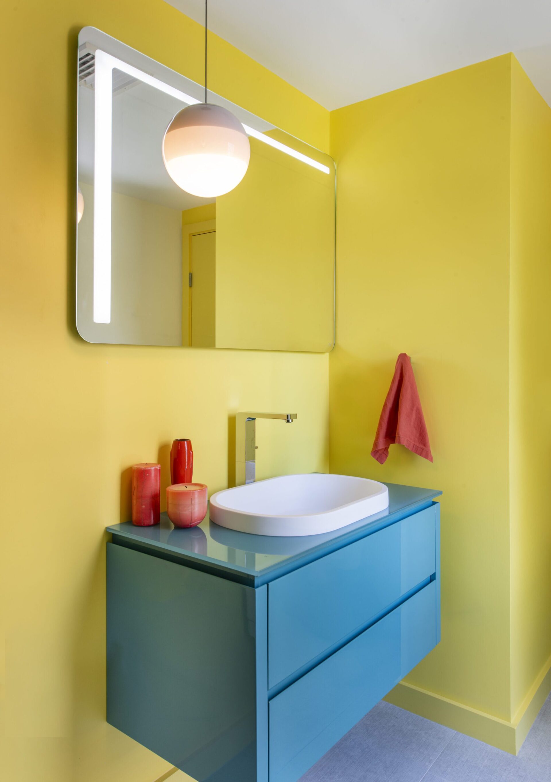 Interior of a house in Quincy, MA, designed by Ruhl Jahnes, showing a bathroom with vivid yellow walls, a floating teal vanity, a white vessel sink, a globe pendant light, and an illuminated mirror.