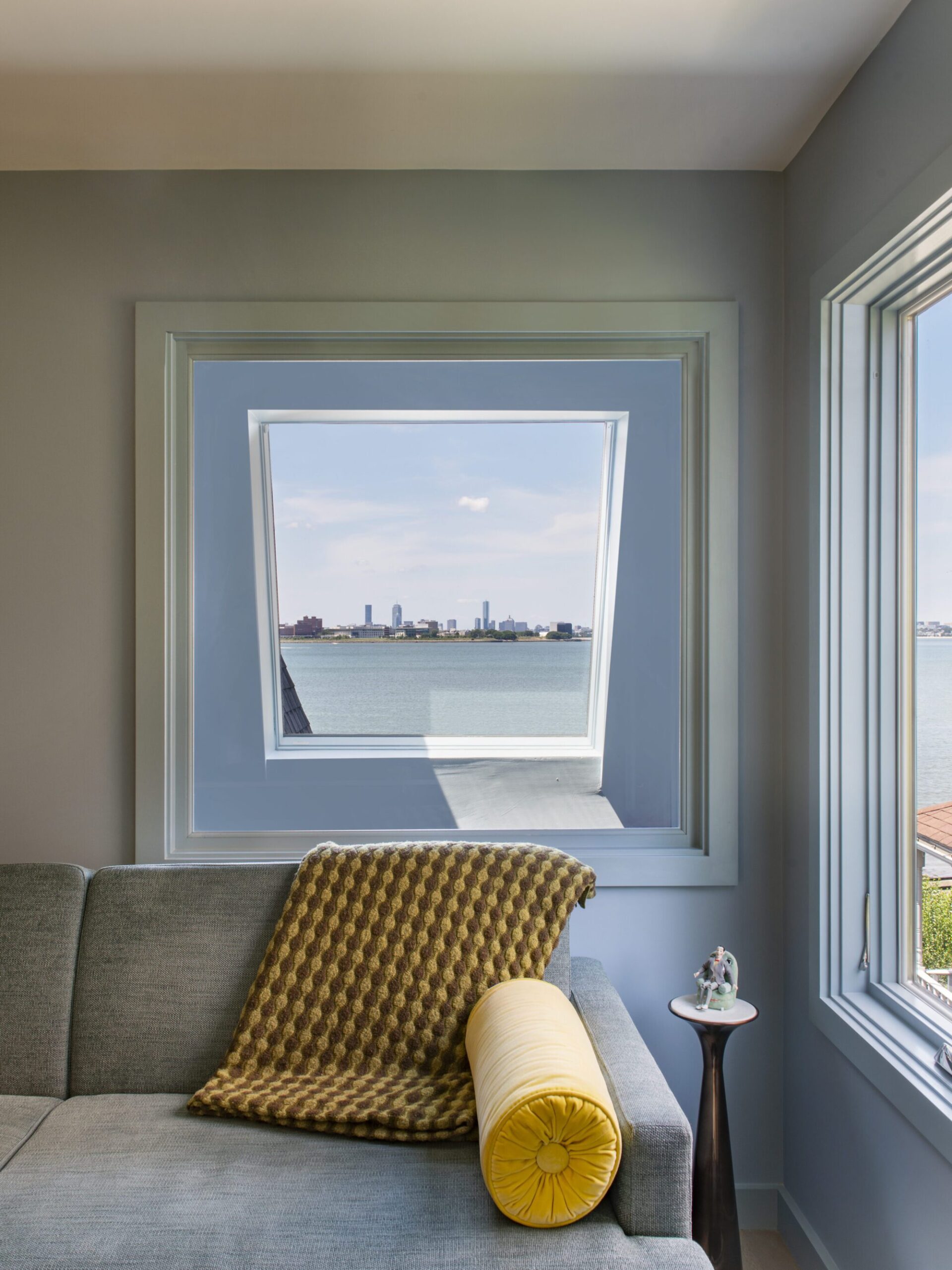 Interior of a Quincy, MA, home designed by Ruhl Jahnes, showing the primary bedroom bay window seating area with a built-in sofa, yellow bolster pillow, blue walls, and a framed view of Boston Harbor and the city skyline beyond.