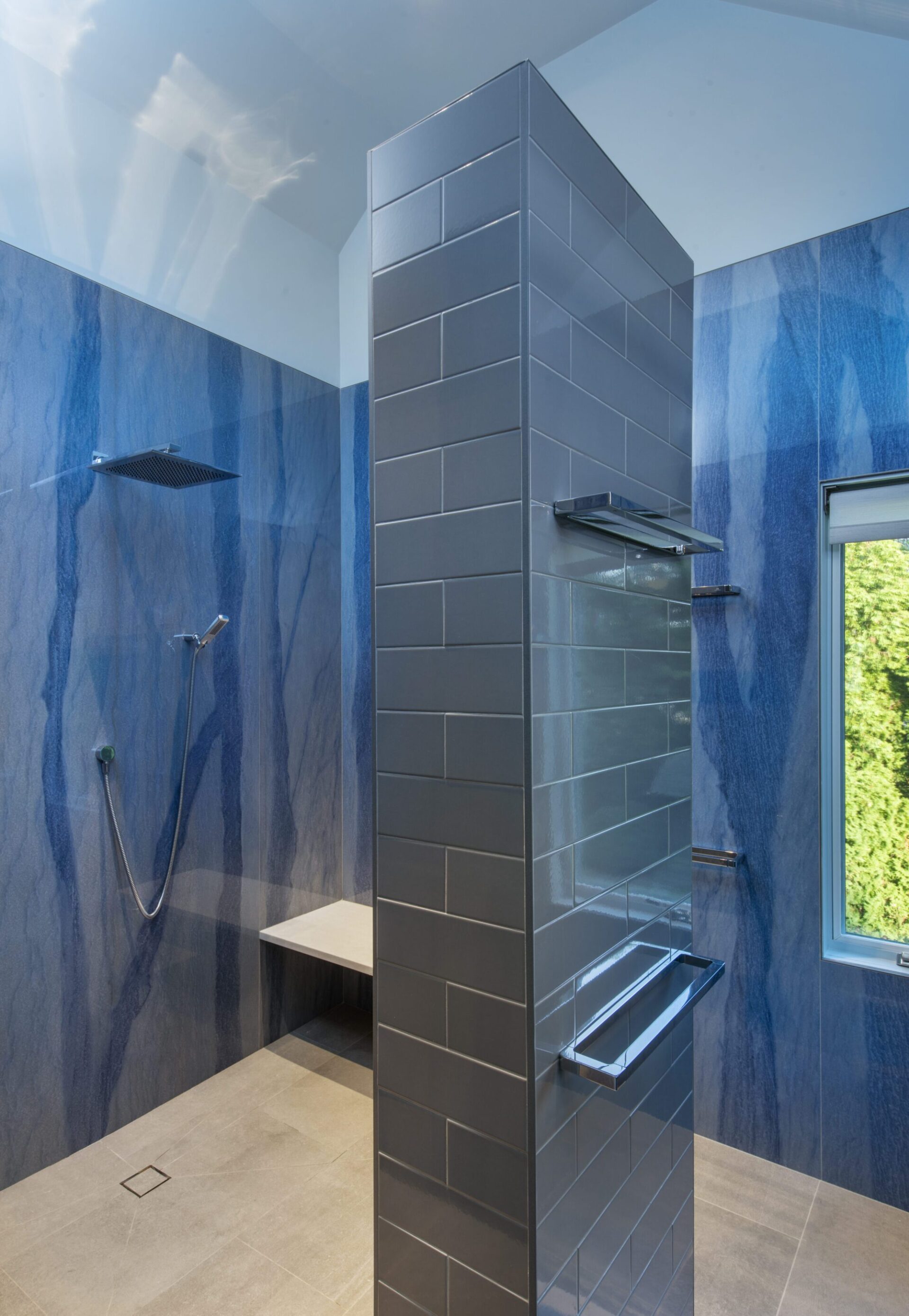 Interior of a house in Quincy, MA, designed by Ruhl Jahnes, showing the primary bathroom with polished cobalt blue quartz wall slabs, a gray tile curbless shower with a built-in bench, rain shower head, and a window overlooking the yard.
