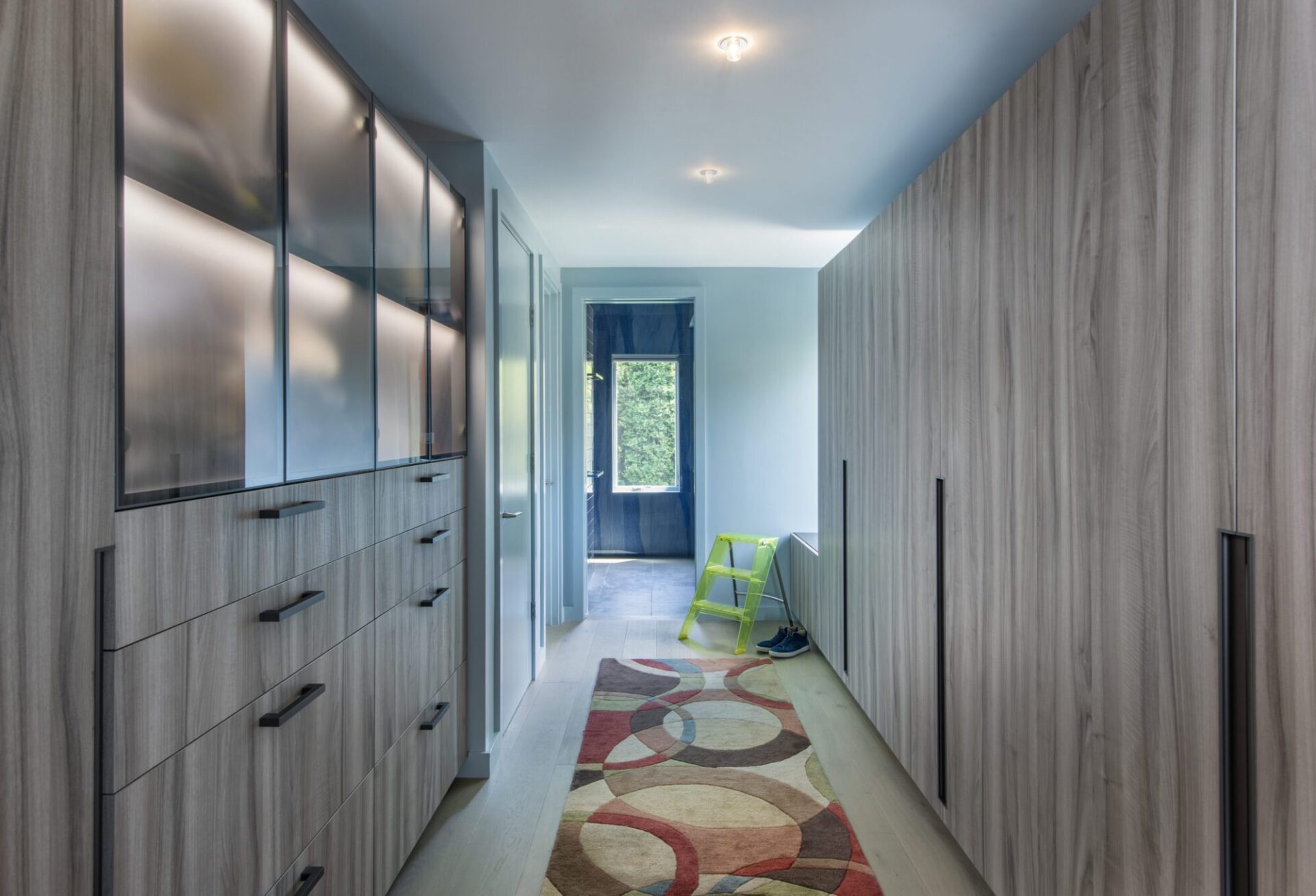 Interior of a Quincy, MA, house designed by Ruhl Jahnes, showing the primary suite dressing corridor with floor-to-ceiling wood-grain cabinetry, a colorful area rug, and a view through to the blue front door beyond.