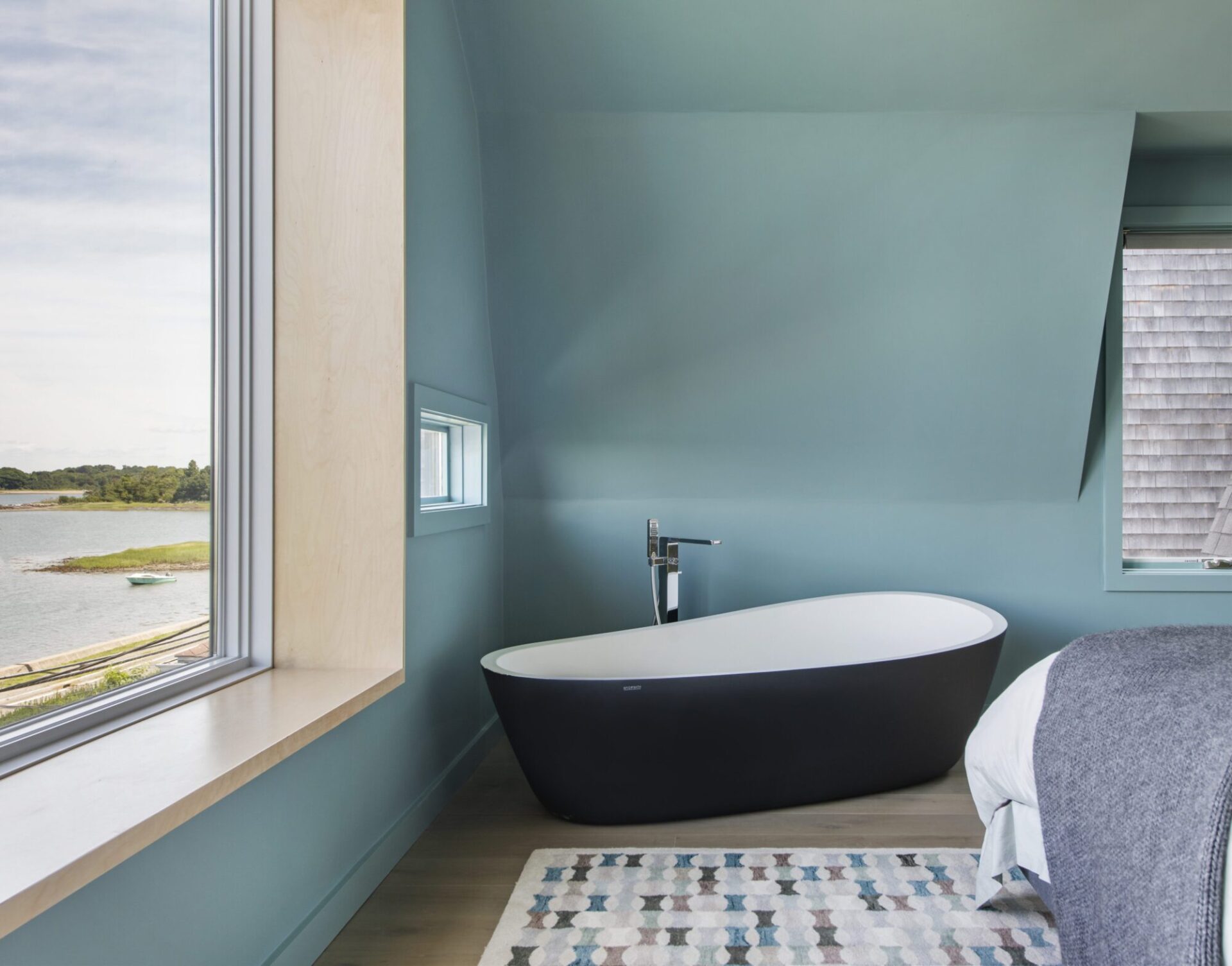 Interior of a house in Quincy, MA, designed by Ruhl Jahnes, showing the primary bathroom with a black and white freestanding soaking tub, blue-gray walls, large windows overlooking the harbor and barrier island landscape, and a mosaic tile bath mat.