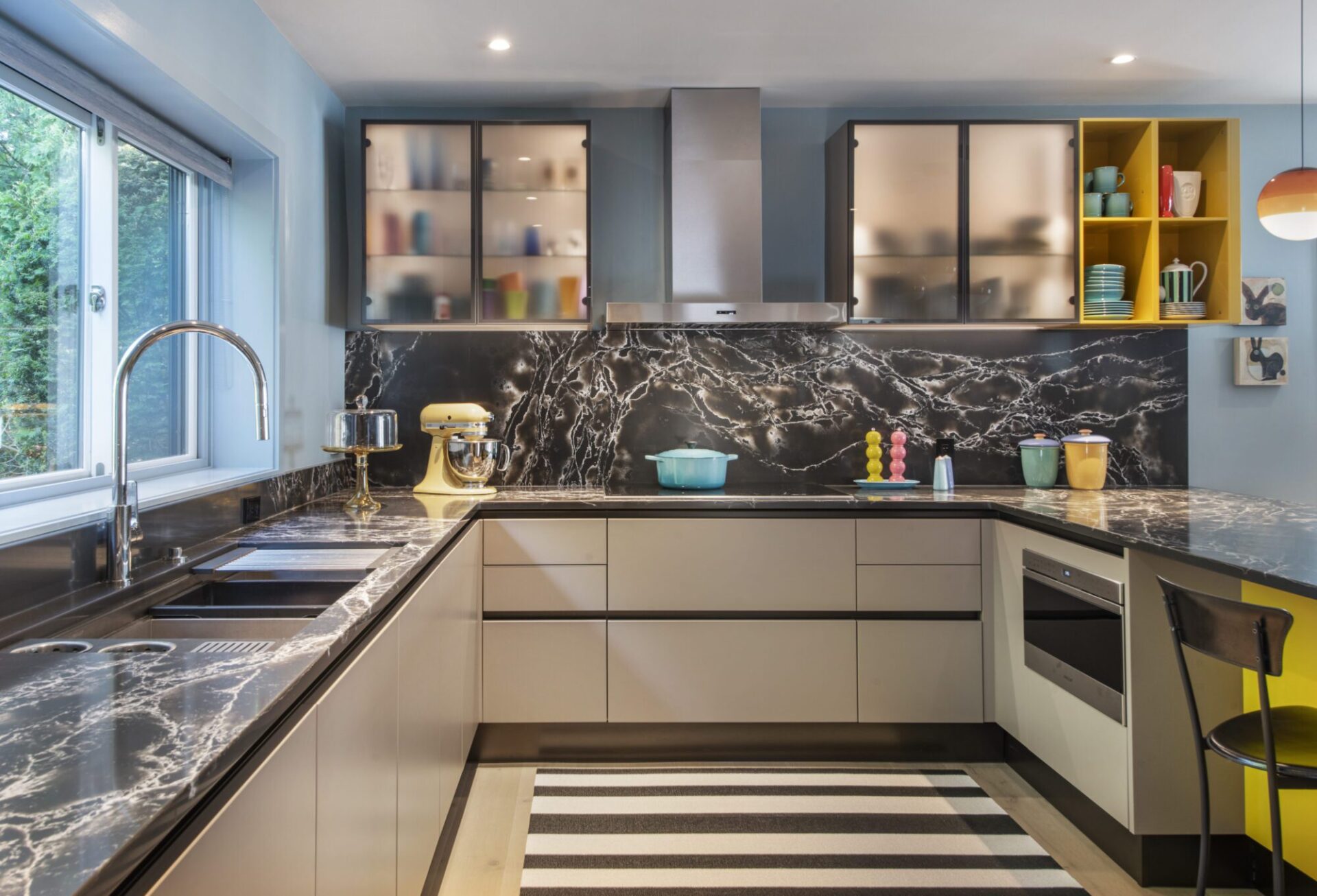 Interior of a house in Quincy, MA, designed by Ruhl Jahnes, showing the kitchen perimeter with black veined quartz countertops and backsplash, white flush cabinetry, glass-front upper cabinets, a stainless steel hood, and yellow open shelving.