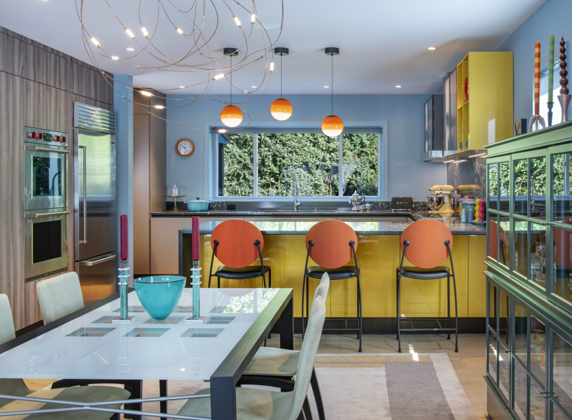 Interior of a Quincy, MA, home designed by Ruhl Jahnes, showing the open kitchen and dining area with a white dining table, orange barstools at the yellow kitchen island, orange globe pendant lights, blue-gray walls, and a sculptural ceiling light fixture.