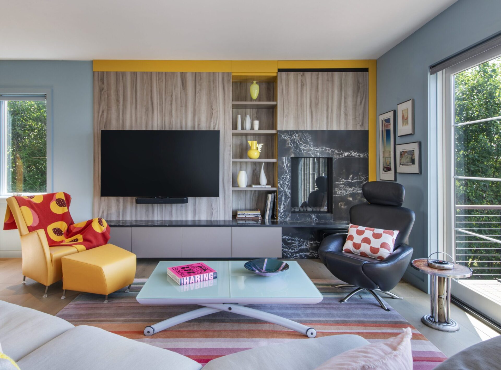 Interior of a house in Quincy, MA, designed by Ruhl Jahnes, showing the living room with a wood-grain built-in entertainment wall, black quartz fireplace surround, yellow accent trim, colorful lounge chairs, a white coffee table, and a sliding glass door to the deck.