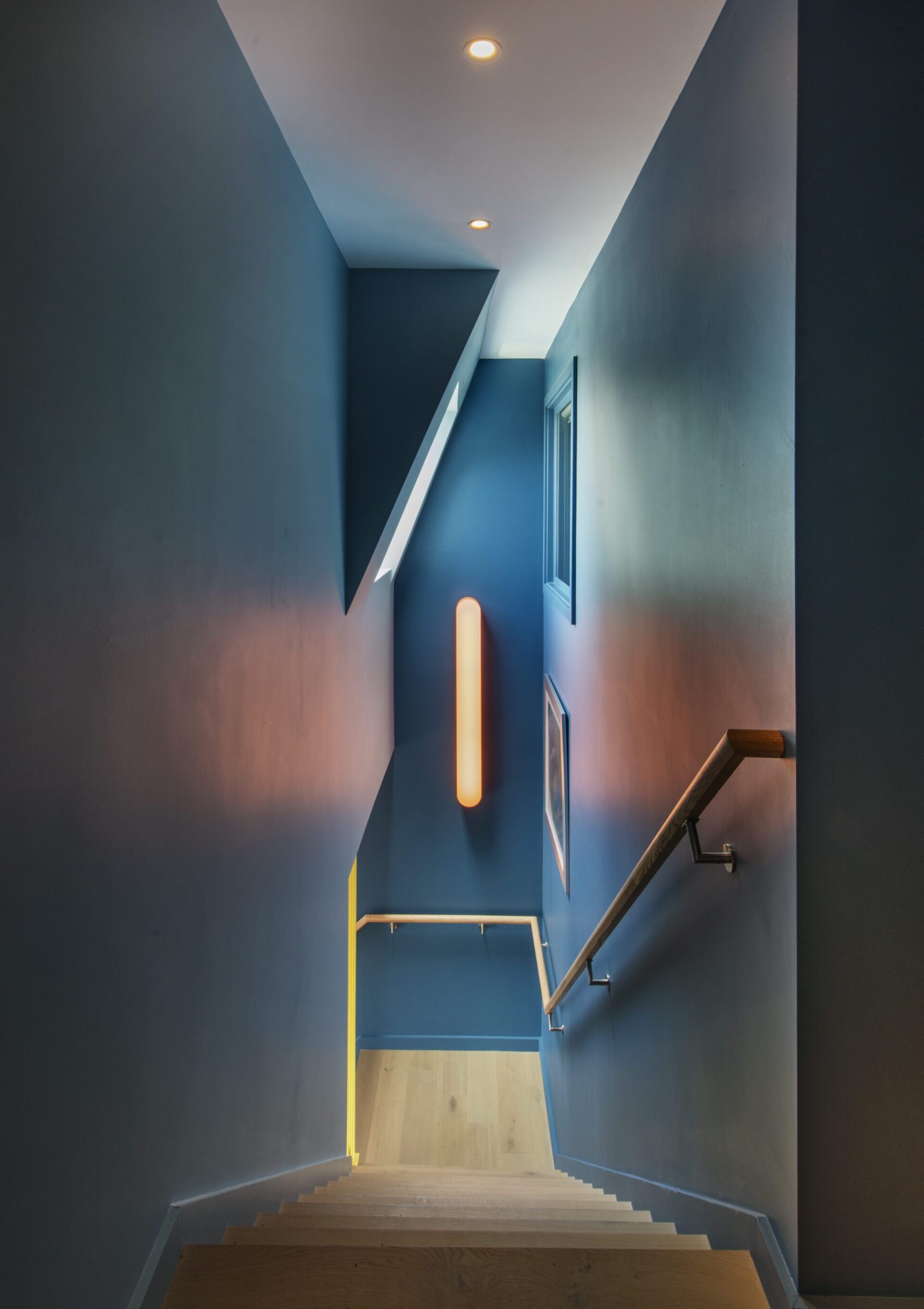 Interior of a Quincy, MA, house designed by Ruhl Jahnes, showing the staircase with deep blue-gray walls, a yellow accent handrail, a wall sconce, and warm wood treads leading down to the entry level.