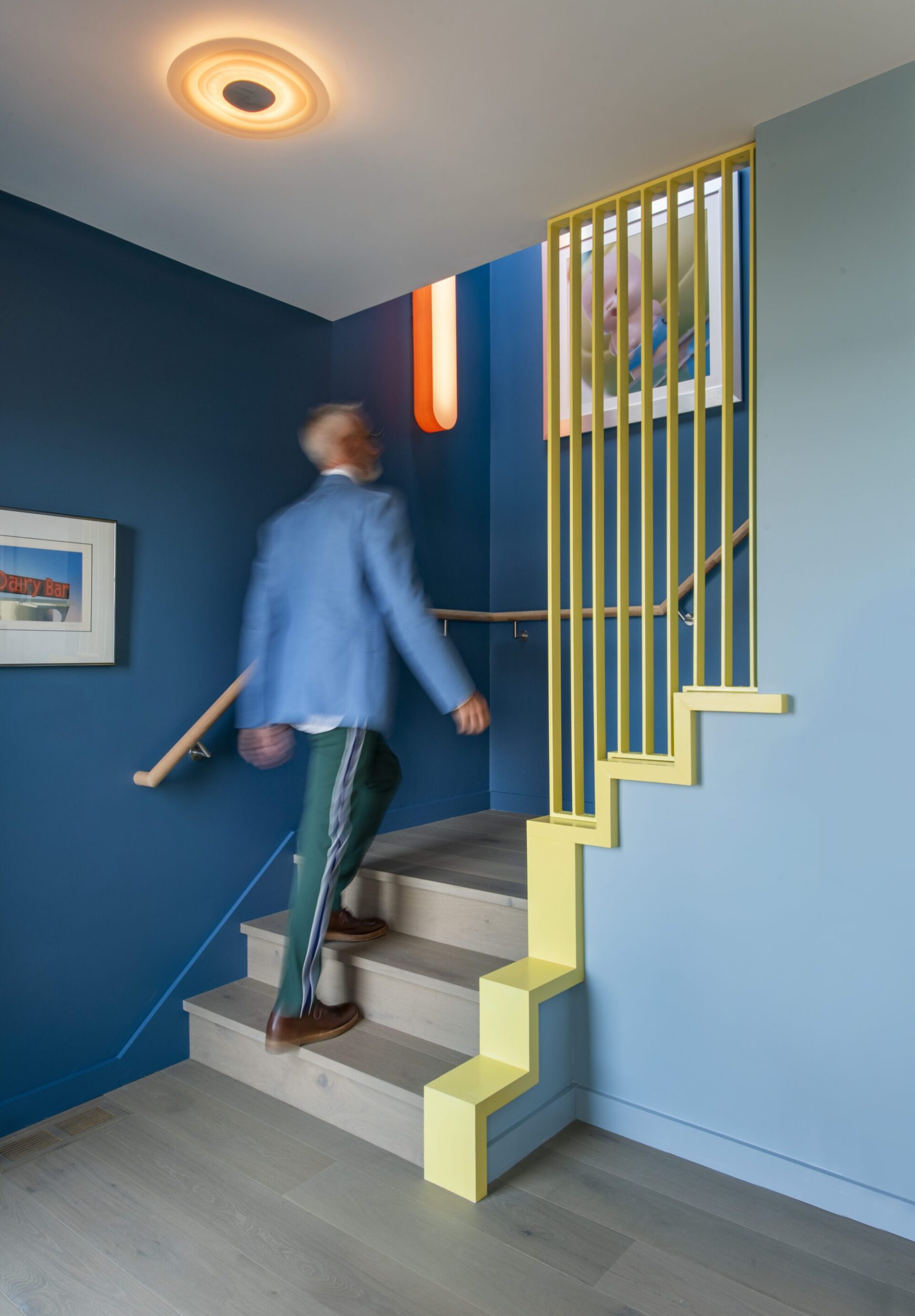 Interior of a house in Quincy, MA, designed by Ruhl Jahnes, showing the entry stair with vibrant yellow trim, a slatted steel screen railing, blue walls, a circular flush ceiling light, and a figure ascending the stairs.