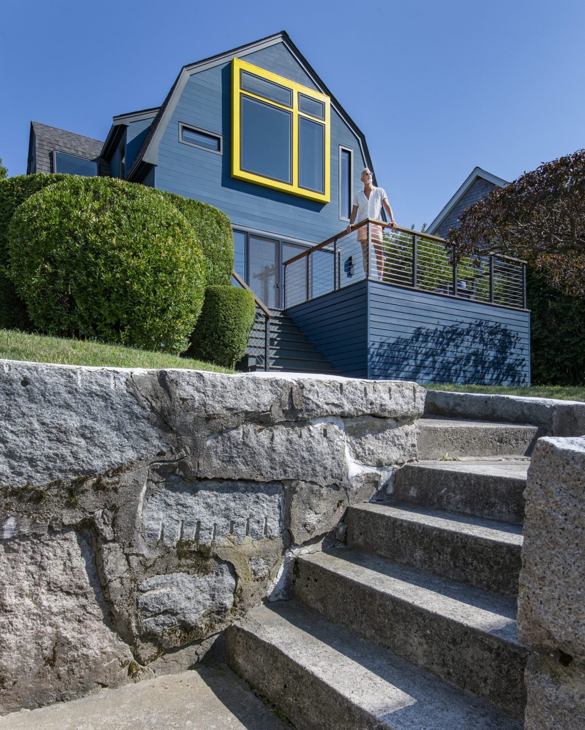 Exterior of a house in Quincy, MA, designed by Ruhl Jahnes, showing the blue facade with yellow bay window surround, harbor-side deck with cable railings, granite block retaining wall, and stone steps leading up from the street.