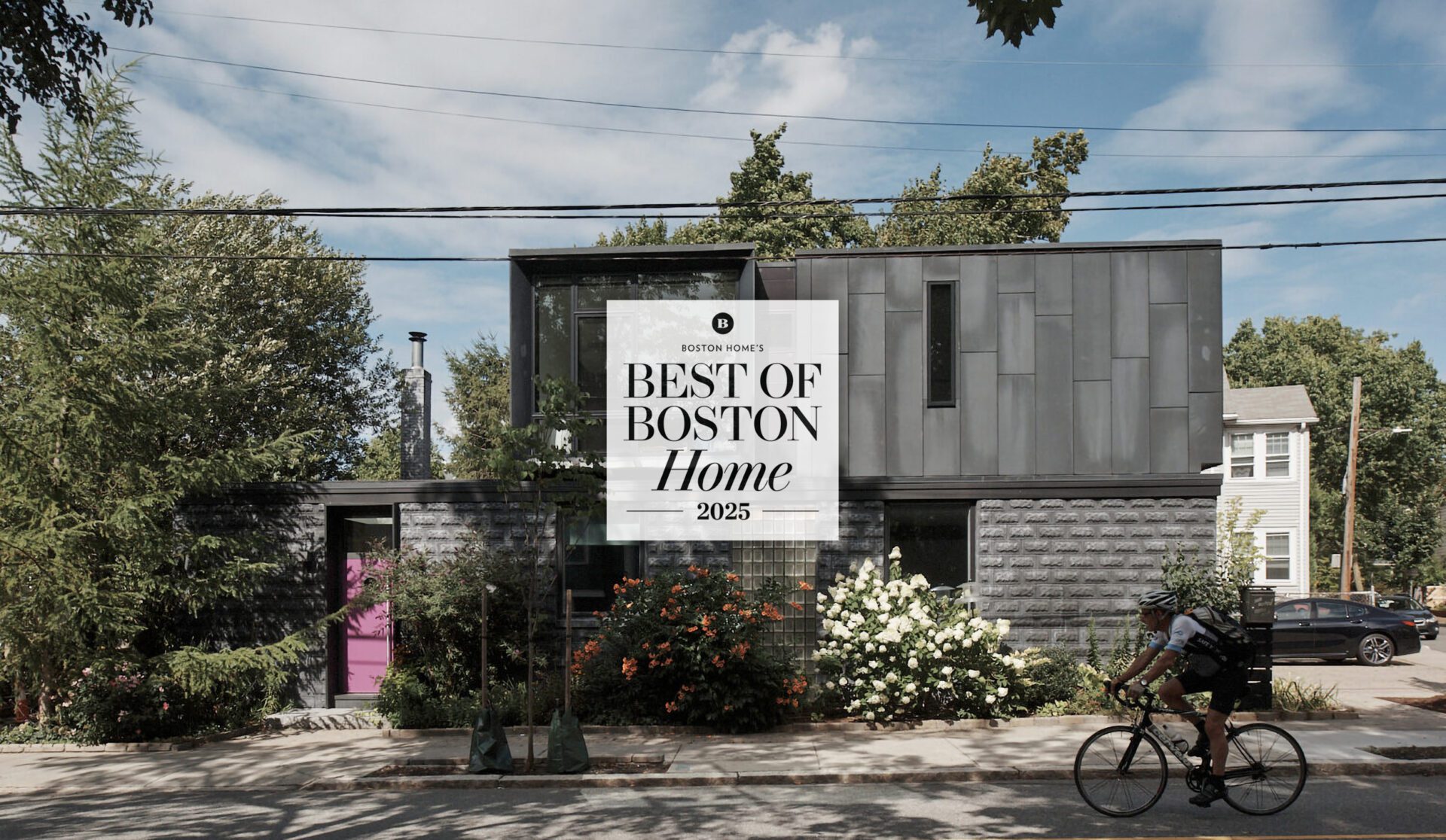 Exterior of a house in Newton, MA, designed by Ruhl Jahnes, showing the Tudor Modern project featured in Boston Home's Best of Boston Home 2025, with the fiber-cement addition, dark cladding, a pink entry door, summer plantings, and a cyclist passing on the street.